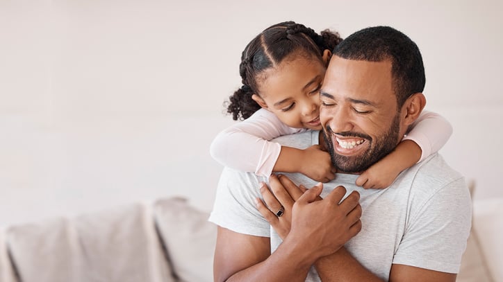 A loving moment between a man and a young child. The child is affectionately hugging the man from behind, with their arms wrapped around his neck and their head resting on his shoulder. Both appear to be smiling and enjoying the embrace.